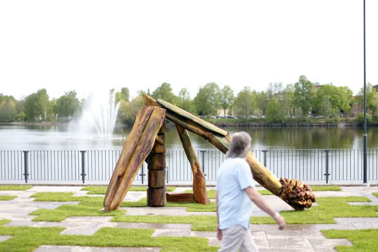 Kuvassa näkymä joen rannasta, jossa puusta tehty veistos, jota ohikulkeva henkilö katsoo.