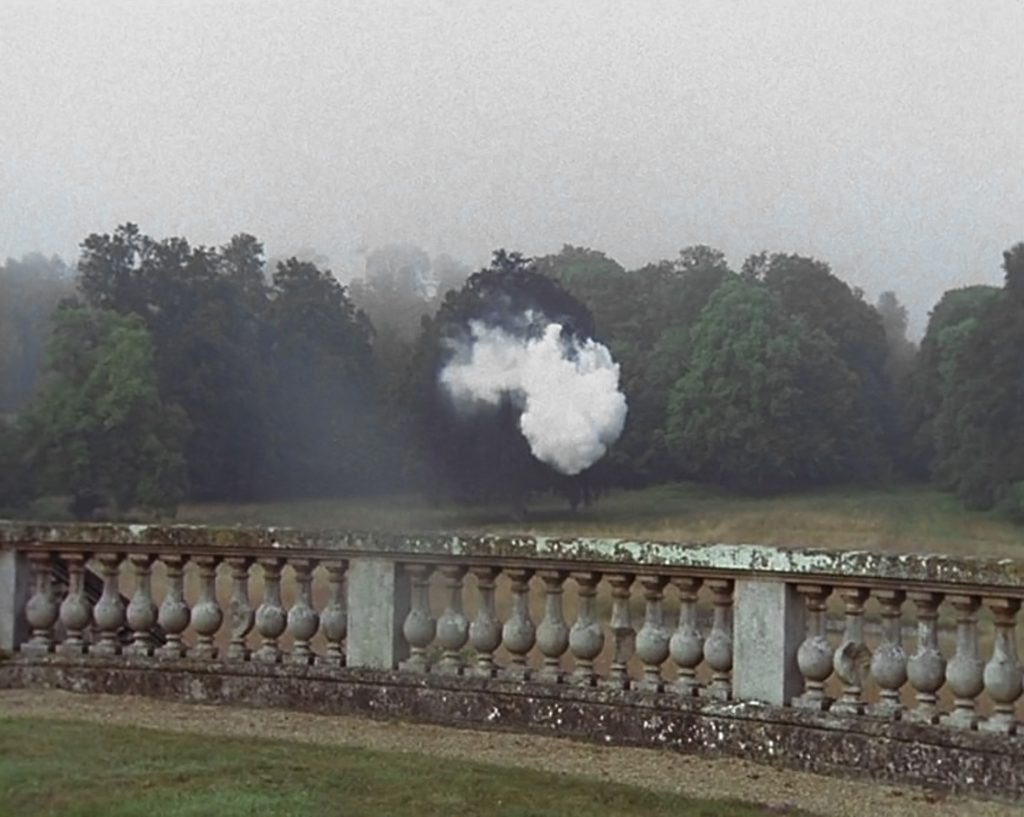Sumuinen maisema, jossa etualalla kivinen kaide, taustalla nurmikko ja tiheä puusto. Puiden lomassa leijuu epätavallinen valkoinen pilvimäinen muodostelma.