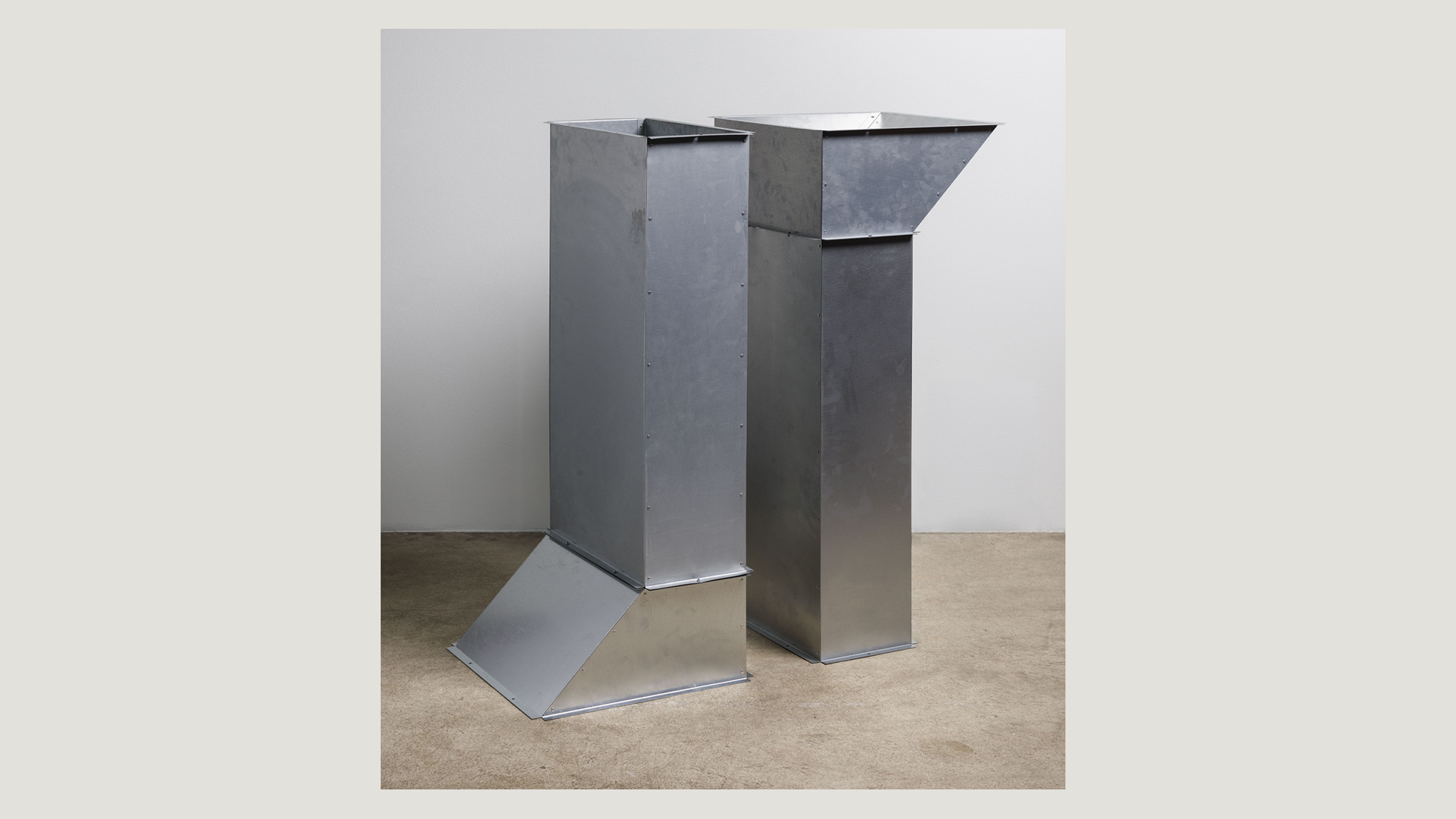 Kaksi metallista geometristä rakennetta betonilattialla vaaleaa seinää vasten.